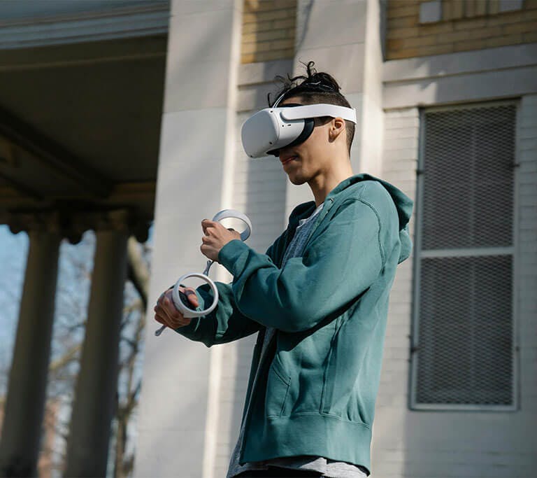 A person wearing a sage zip-up hoodie, a grey shirt, and a virtual reality (VR) headset stands outside of a house on a sunny day and plays a VR game.