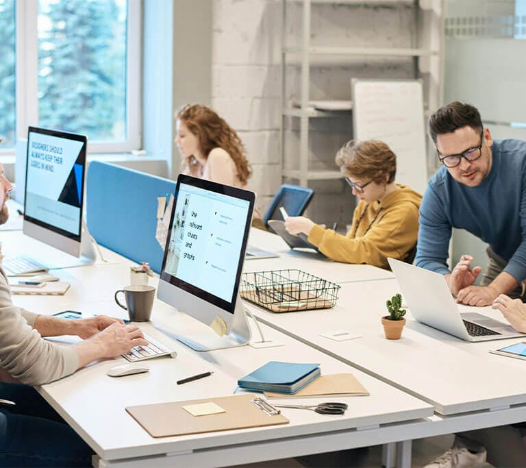 People working together in a modern office with laptops and desktop computers; one person appears to be assisting a colleague while others focus on their screens.
