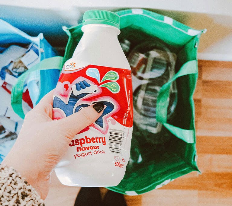 A person sorts an empty bottle of “raspberry flavour yogurt drink” into a green recycling bag as part of their sustainability goals.