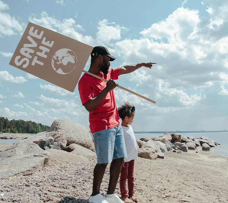 A father carrying a sign that says “Save the Earth” points at the ocean while his child looks at the water on a beautiful day.