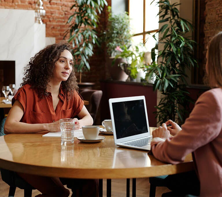 One person wearing a mauve blazer uses a laptop across the table from another person whose hands are resting on a stack of papers in cafe decorated with tropical plants.