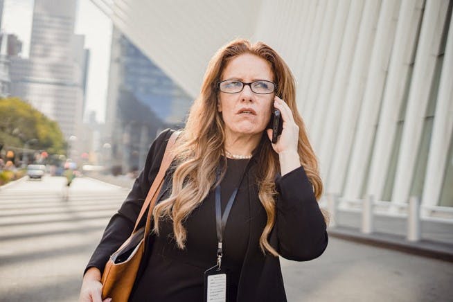 A woman with a lanyard and a purse is walking through a city holding a cell phone to her ear with a disgruntled expression on her face.