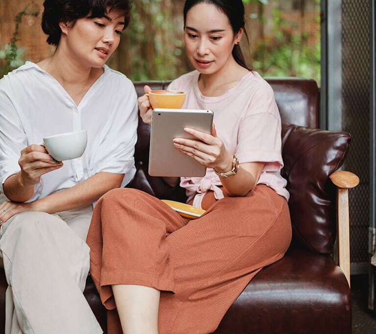 Two people sitting on a leather sofa in a cozy café, holding coffee cups and looking at a tablet together. The setting includes warm lighting and greenery visible in the background, creating a relaxed atmosphere.