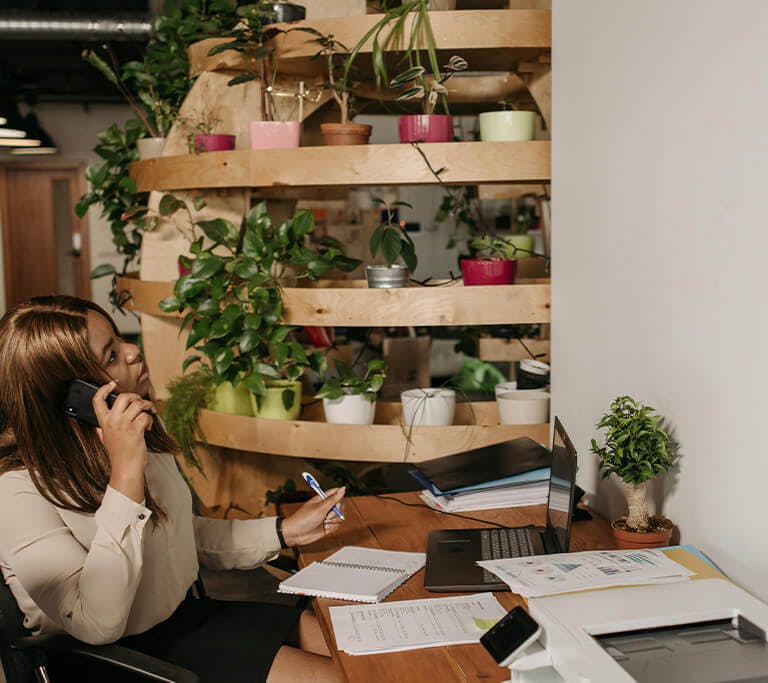 A person sits at a desk in an office, talking on the phone while taking notes and working on a laptop. Papers, folders and a printer are spread across the workspace, and a large wooden plant display filled with potted plants stands behind the desk.