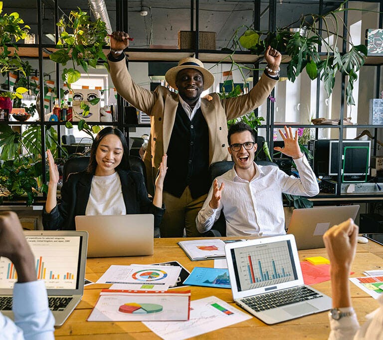 A group of colleagues celebrating success in a modern office with laptops and charts on the table. One person stands with raised arms in excitement while others smile and cheer, surrounded by plants and natural light.