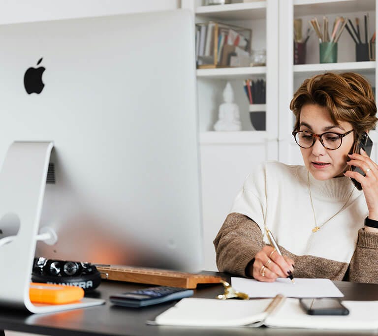 A person sits at a desk working on a large computer monitor while talking on the phone and writing notes on a sheet of paper. The desk is covered with office items, including a notebook, smartphone, glasses and external hard drive, with shelves of books a