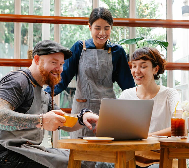 Three people wearing café aprons gather around a laptop at a small wooden table, smiling while reviewing something on the screen. A drink and a small vase of flowers sit on the table beside the laptop, with large windows and greenery in the background.