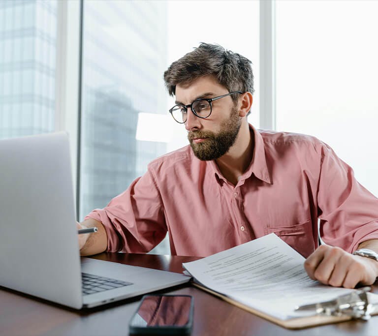 A person wearing glasses sits at a desk in front of a laptop, reviewing documents on a clipboard while holding a pen. A smartphone lies on the table nearby. Large office windows reveal tall buildings in the background.