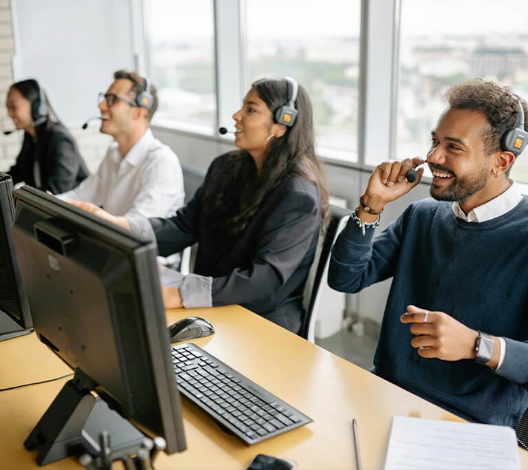 A group of customer support agents sits in a row at desks, wearing headsets and working at desktop computers. One person in the foreground smiles while speaking into a headset. Large windows behind them show a cityscape in the distance.