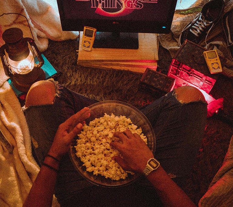 A person sits in a cozy blanket fort holding a bowl of popcorn while streaming on a small display surrounded by lantern light and string lights.