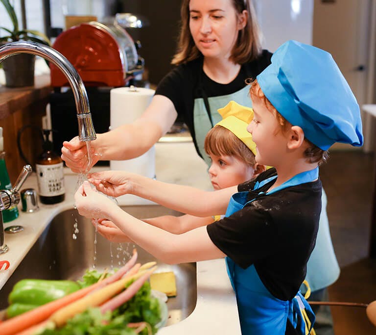 A mother and two children wearing chef hats and aprons wash their hands in the kitchen sink, with carrots and celery nearby.