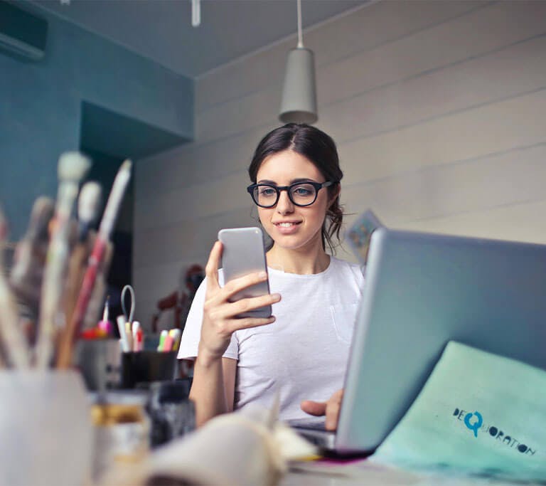 A person wearing glasses and a white T-shirt smiles at the phone while typing on a laptop in an art studio.