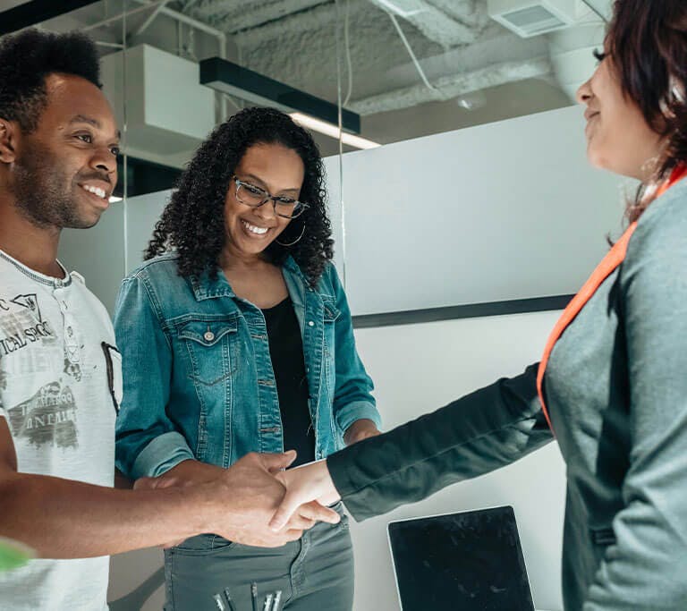 Three people standing in an office setting. Two of them are smiling and shaking hands across a desk, while the third person looks on with a smile. A laptop and pens are visible on the desk in front of them.