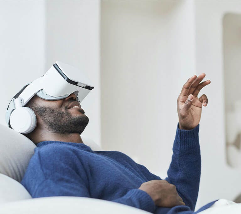 A person wearing a blue sweater and a white virtual reality gaming headset smiles and reaches upward with one hand while resting the other on their stomach as they recline in a white chair.