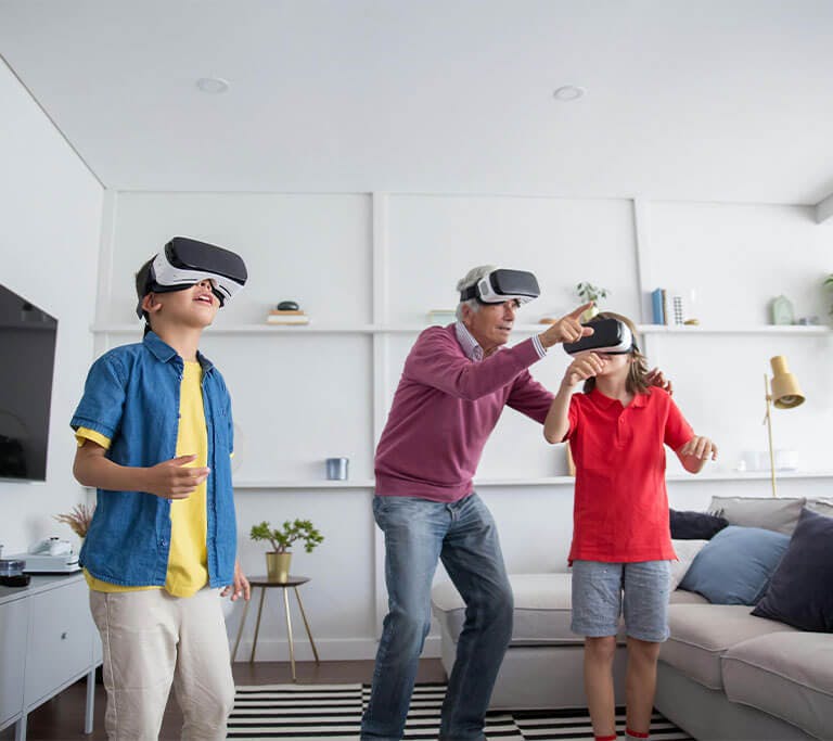 A grandparent and two grandchildren wear VR headsets in a living room while pointing and interacting with an off-screen game.