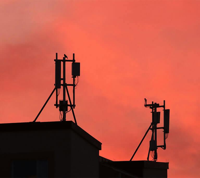 Cellular antennas mounted on a rooftop, silhouetted against a vivid orange sunset sky.