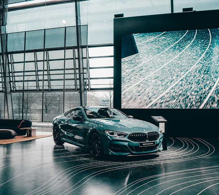 A sleek car displayed in a modern showroom with large glass windows and a giant digital display showing abstract track lines. The polished floor features curved white patterns, and there’s contemporary seating in the background, creating a high-end, futur