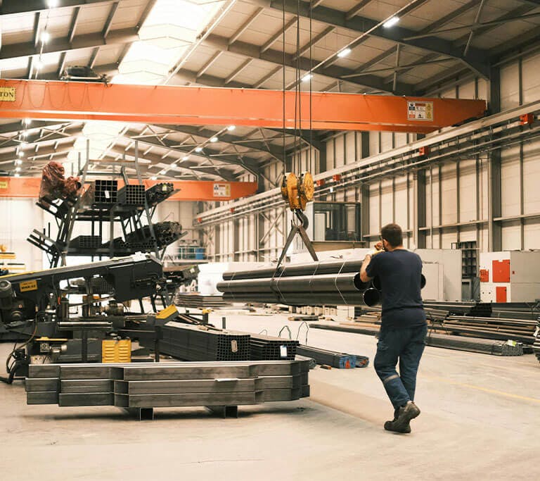 A person working inside a large industrial warehouse, guiding metal pipes being lifted by an overhead crane. The facility is spacious with high ceilings, steel beams, and organized stacks of metal materials and machinery.