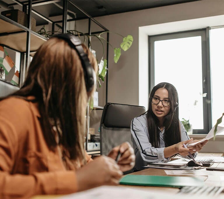 Two people wearing headsets sit at a shared office desk, with one holding papers and talking to the other near a bright window and indoor plants.