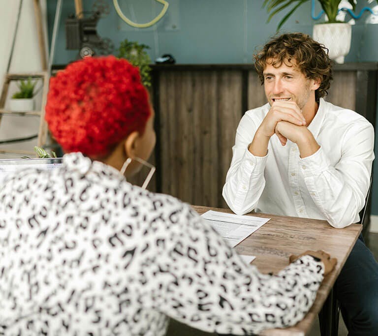 Two people sitting across from each other at a desk having a friendly conversation, with papers and a laptop in front of them in a modern office setting.