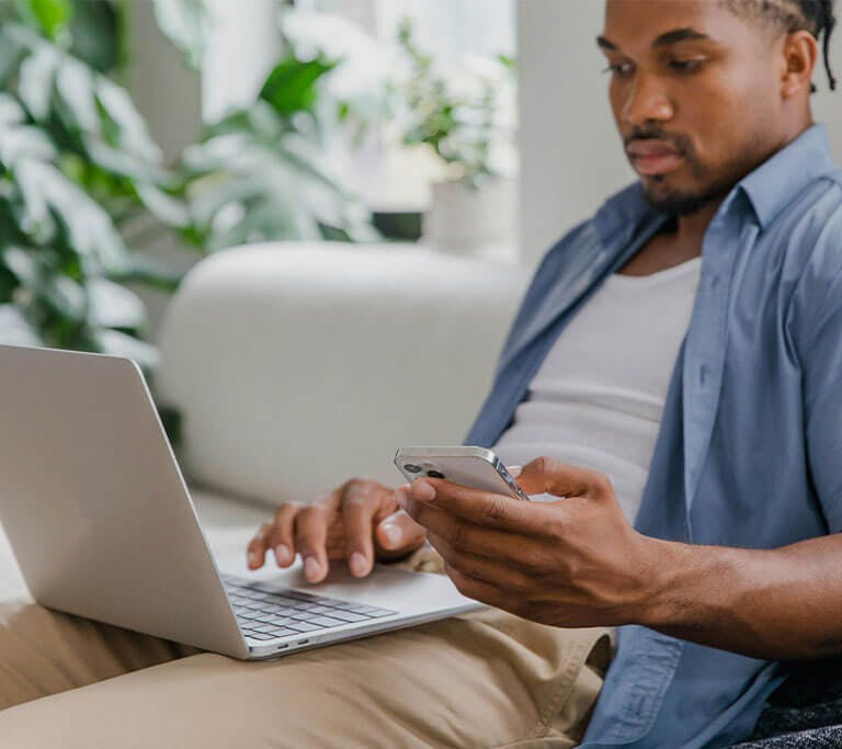 Person sits on a couch using a laptop while checking a smartphone, with plants in the background.