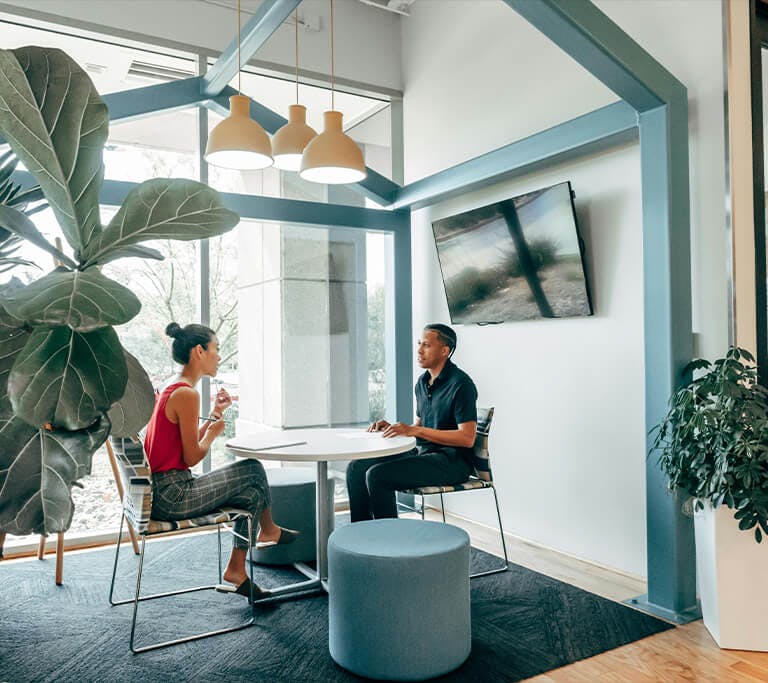 Two people sitting across from each other at a round table in a modern office space with large windows and indoor plants. The area features hanging lights, a mounted TV, and a calm, open atmosphere suitable for a meeting or discussion.