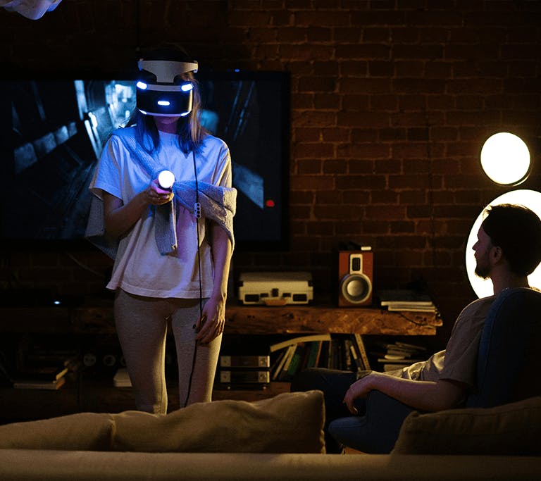 A couple in a brick room that’s dimly lit with a modern, minimalist lamp try out a VR game together, with one person standing, wearing a headset and holding a joystick and the other person sits in a chair and watches.