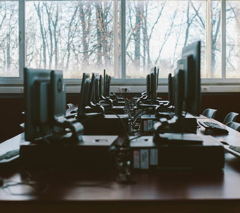 Several computers sit on a desk close together next to large windows.