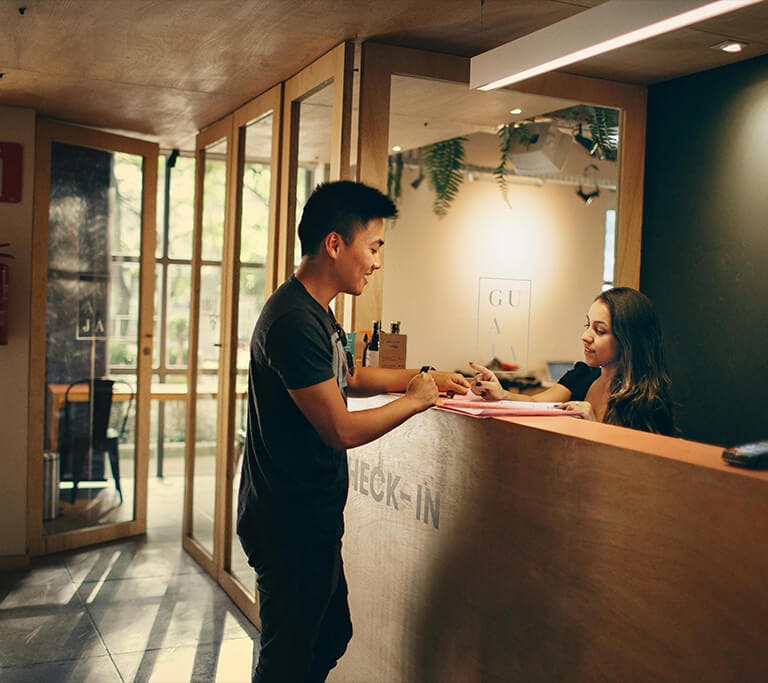 A person standing at a reception desk talking with a receptionist inside a modern building. The receptionist hands over a document while the person smiles and signs it, suggesting a check-in or registration process.