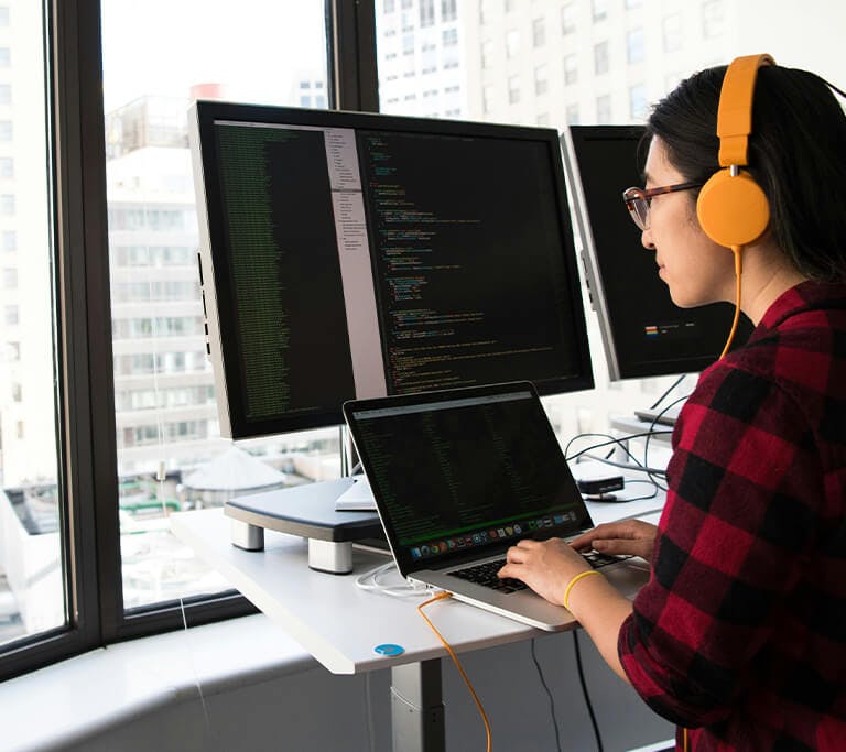 A person wearing bright headphones works at a standing desk with a laptop connected to two large monitors displaying lines of code. Sunlight comes through tall windows overlooking a cityscape of office buildings.