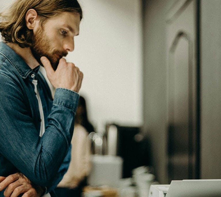 Person stands near a printer with a thoughtful expression, appearing to troubleshoot a printing issue.