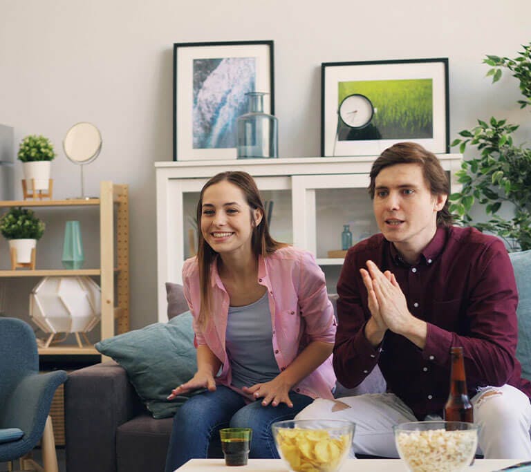 Two people sit on a couch watching TV, with bowls of chips and popcorn on the coffee table in a bright living room.
