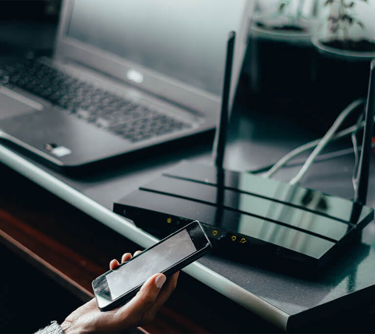 Person sitting at desk holding smart phone, laptop is open, connecting devices to router