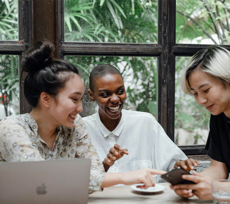 Three friends smile and laugh at a table in front of large windows as they point at a cell phone.