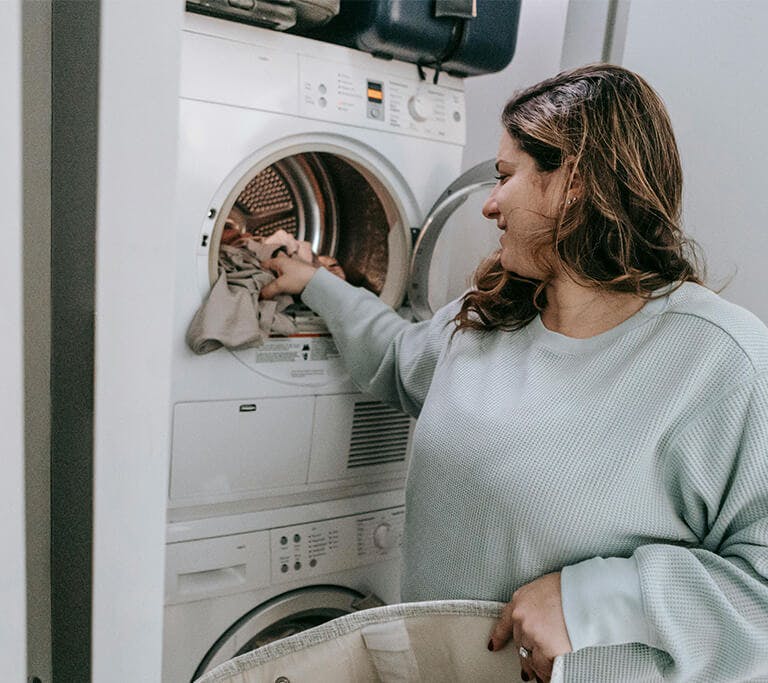 A person with medium-length brown hair smiles as they load clothes into a washing machine.