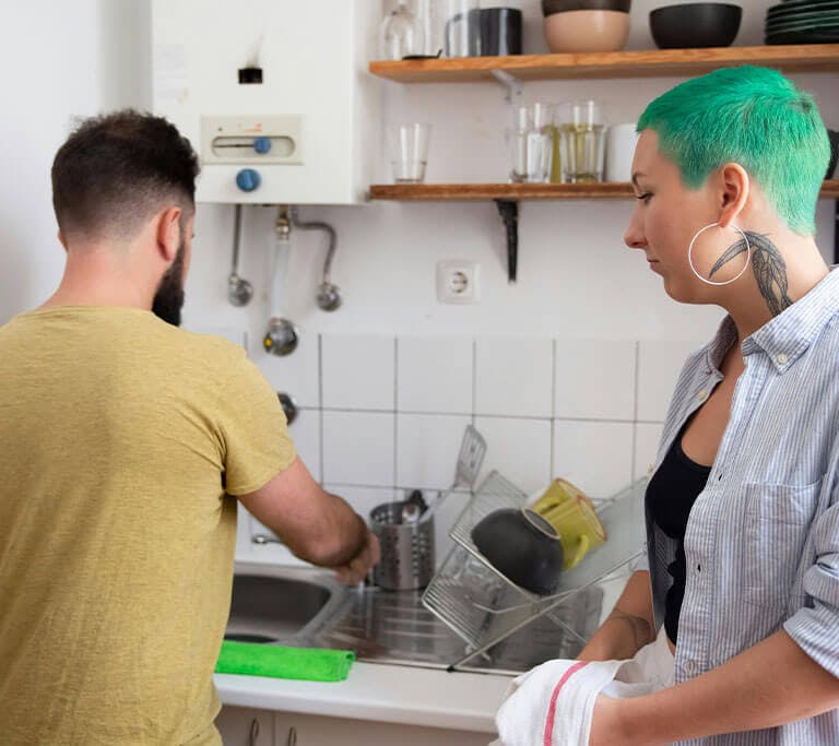 A couple washes dishes in the sink using water that's been heated by the water heater in the background.