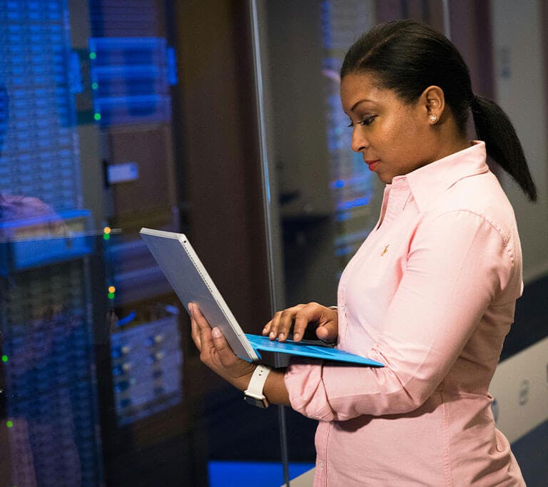 A person stands in a server room working on a laptop. They are focused on the screen, surrounded by illuminated server racks behind a glass panel, indicating they are likely performing IT or network-related tasks.