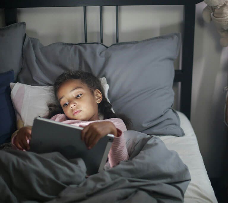 A child wearing pink pajamas lies in bed and looks at a tablet.