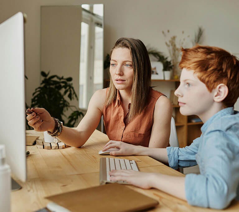A mother and child sit at a desk in a home office while the mother points at the screen with a pen and the child uses a keyboard and mouse while looking at the display.