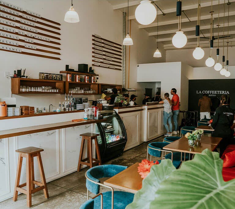 A cozy café with warm LED lighting and modern décor, featuring a long counter with bar stools, a glass display case with drinks and desserts, and several people ordering at the register. The seating area includes wooden tables with blue chairs and plants.