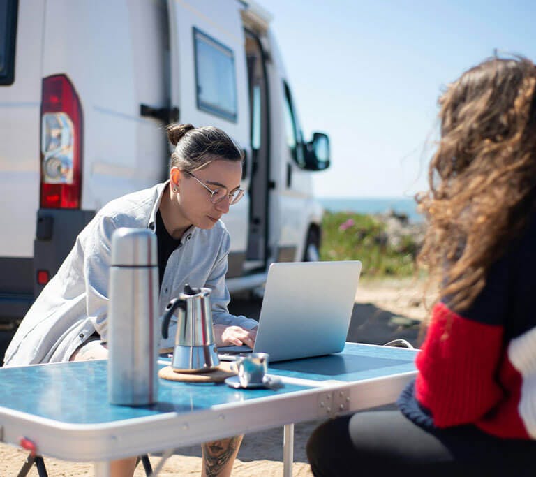 Two people sitting at a folding table outdoors near a camper van, with one using a laptop and a coffee maker and thermos on the table, set against a backdrop of blue sky and ocean.
