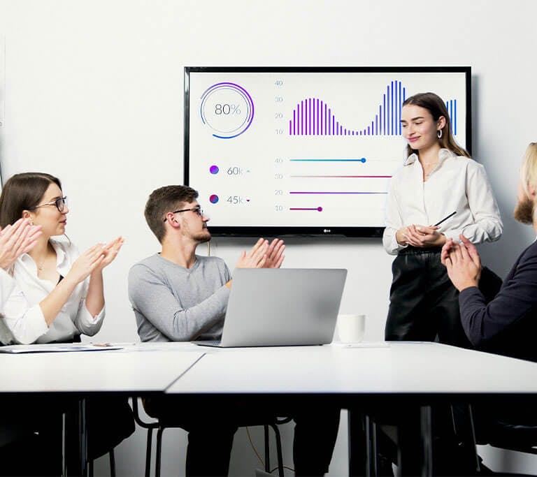 A group of colleagues in a meeting room applauding a presenter standing near a screen displaying colorful charts and graphs. The setting appears professional, with laptops, documents, and a flip chart on the table.