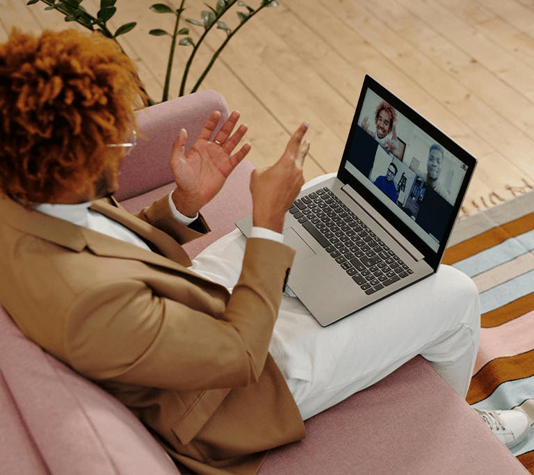 A person sits on a pink couch with a laptop on their lap, participating in a video call with four others visible on the screen. They gesture with their hands while speaking, seated in a bright room with wooden floors and a striped rug nearby.