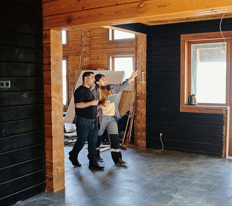 Two people stand in a partially renovated wooden room, one holding a tablet and gesturing toward a wall while they discuss plans near large windows.