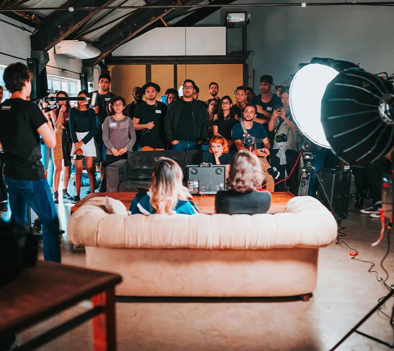 A group of people gathered in a studio during a filming session, with cameras, lighting equipment, and two individuals seated on a couch being filmed while others watch attentively.