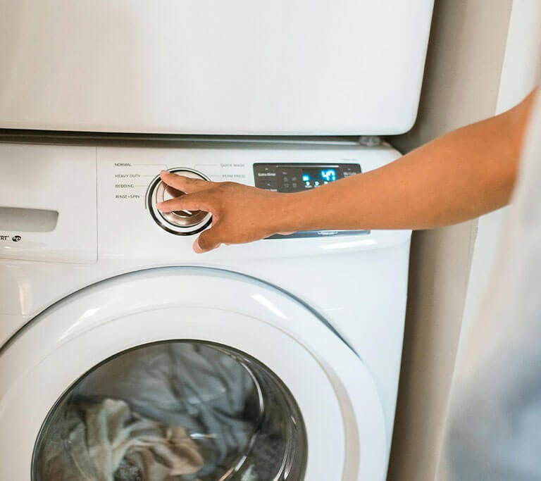 Hand turns the control dial on a front-loading washing machine with laundry visible inside.