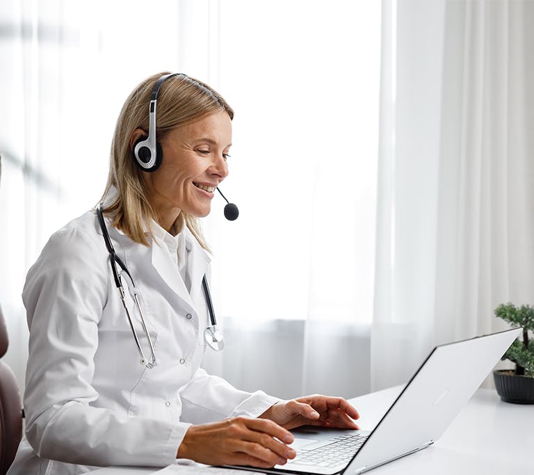 A medical professional sits in a white room at a white desk while wearing a headset as they consult with a patient over a video call.