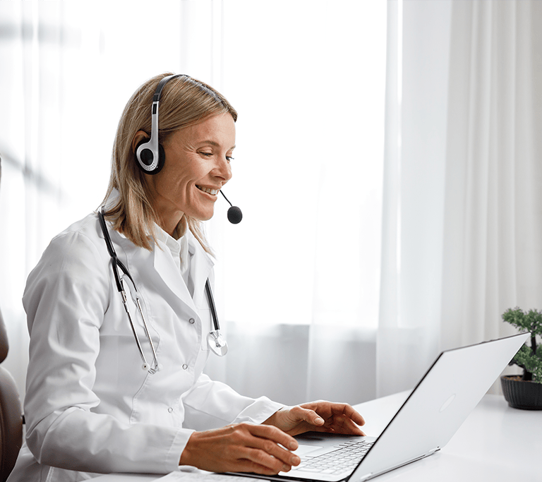 A medical professional sits in a white room at a white desk while wearing a headset as they consult with a patient over a video call.
