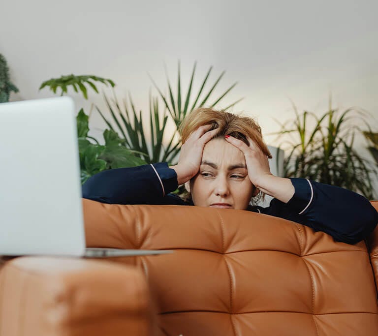A person wearing a navy blue top peers over the back of their brown leather couch with an exasperated expression on their face as they look at their laptop computer that’s sitting on an armrest.