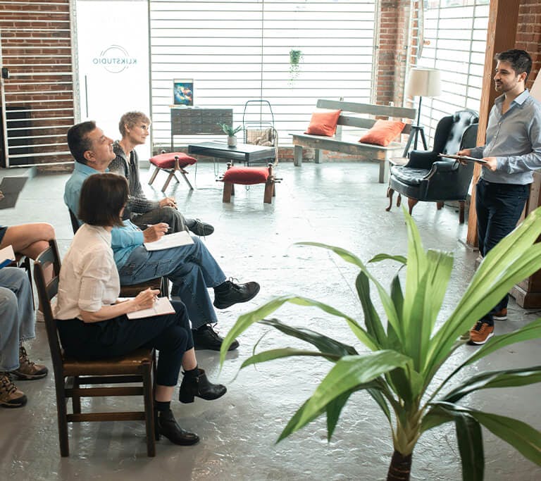 A group of people sit in chairs with notebooks, listening to a presenter at the front of a bright studio space with brick walls, large windows and plants.